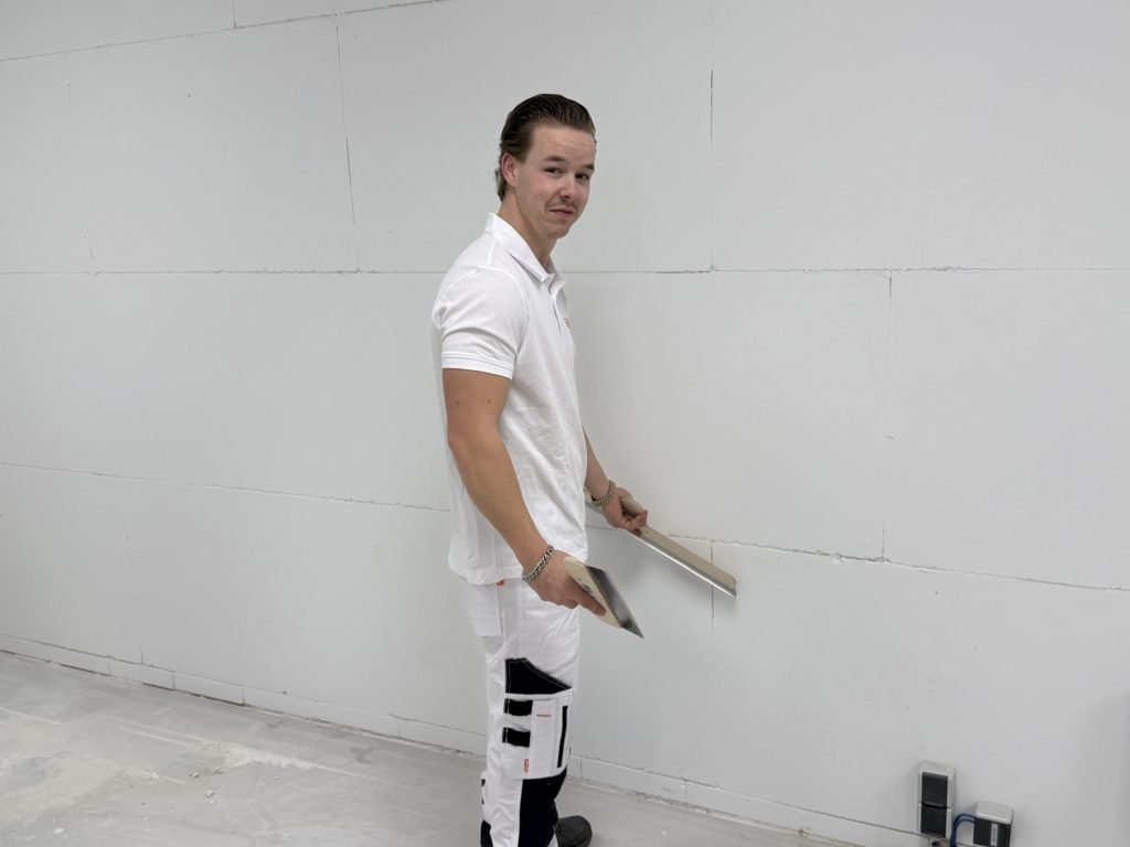 Jonge stukadoor houdt spaan en plakspaan onderaan een wand en kijkt om naar de camera