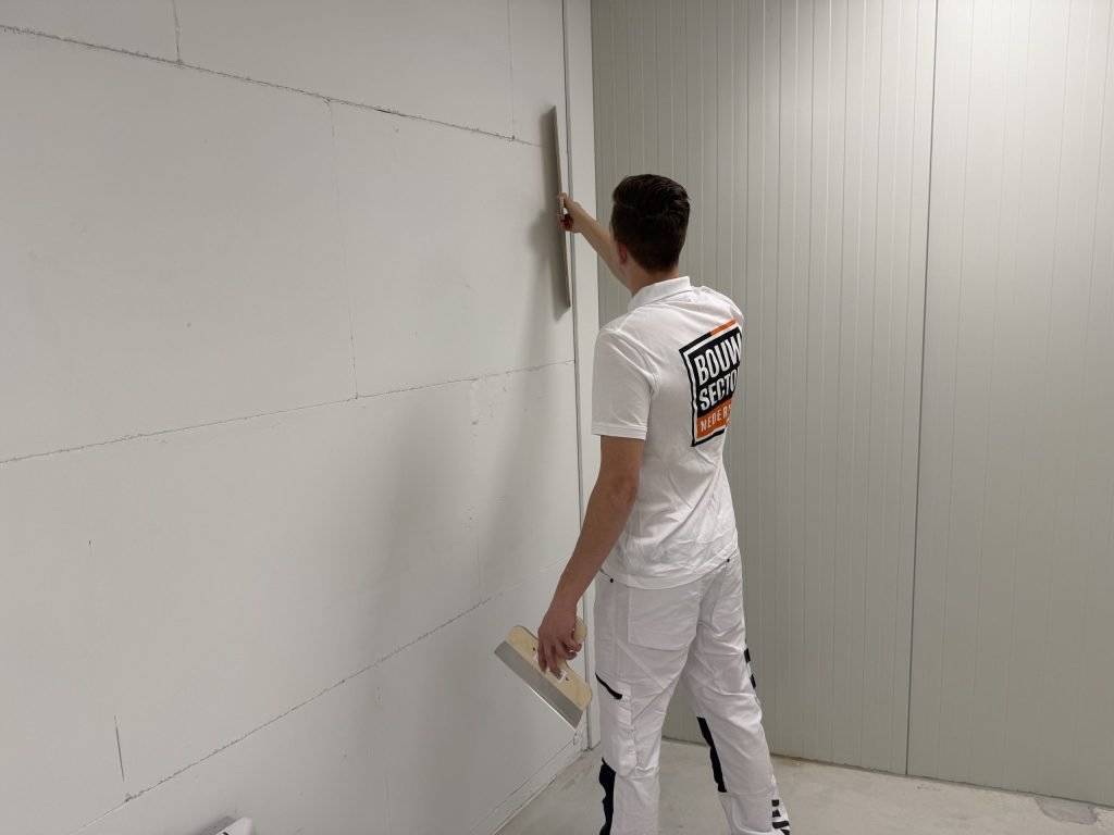 Stukadoor in wit werkpak loopt met de lange spaan langs de naad van een grijze wand
